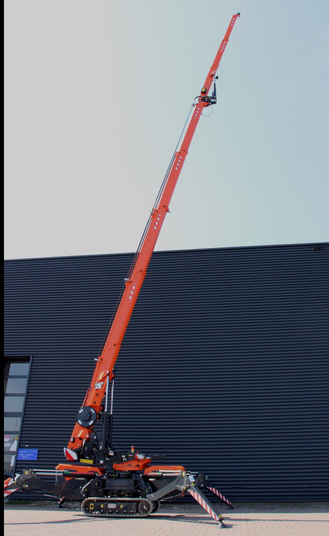 Pomarańczowy dźwig gąsienicowy, wysięgnik w górze, stabilizatory rozłożone, na tle ciemnej hali. Widoczne detale konstrukcji i mechanizmów.