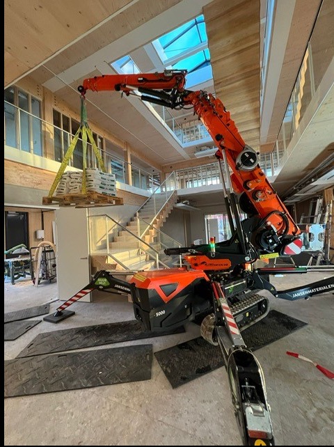 Pomarańczowy mini dźwig gąsienicowy Jansen S5000 podnosi paletę z materiałami wewnątrz budynku z drewnianą konstrukcją i schodami.