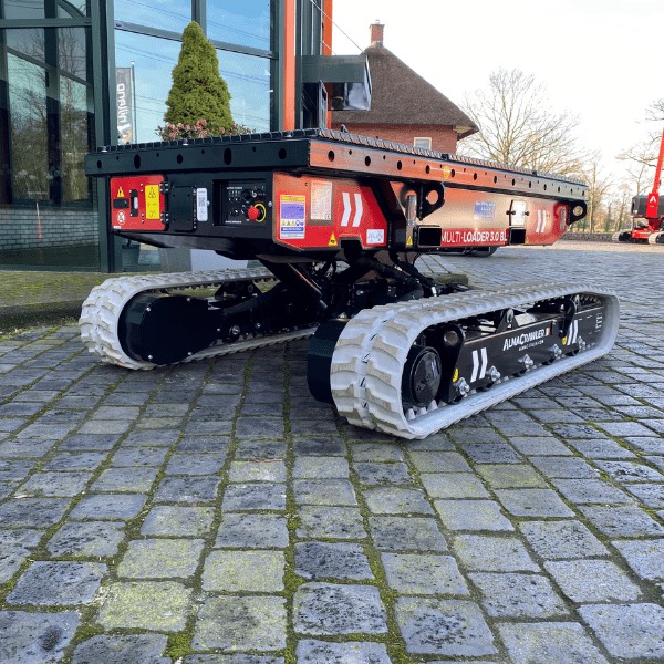 Czerwono-czarny samojezdny mini dźwig gąsienicowy Multi-Loader 3.0 BL na brukowanej nawierzchni. W tle budynek z zielonymi elementami i drzewo.