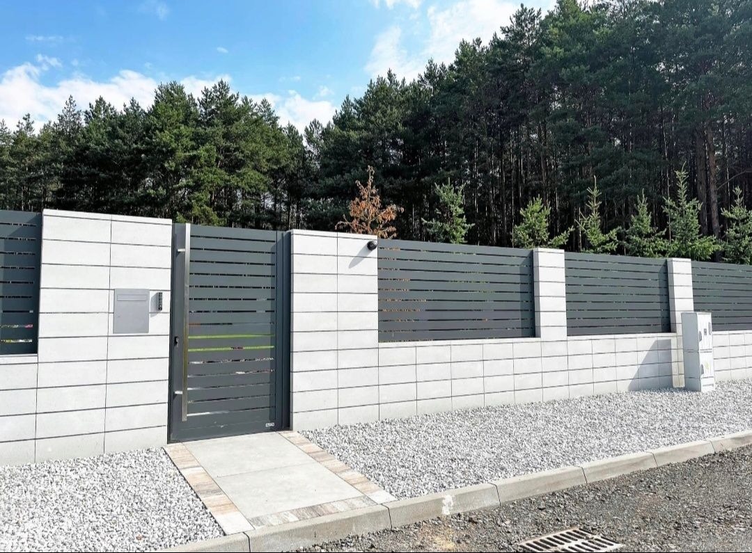 Nowoczesne ogrodzenie betonowe z metalowymi przęsłami i furtką w kolorze grafitowym. Widoczna skrzynka na listy i kamera. Tło stanowi las iglasty.