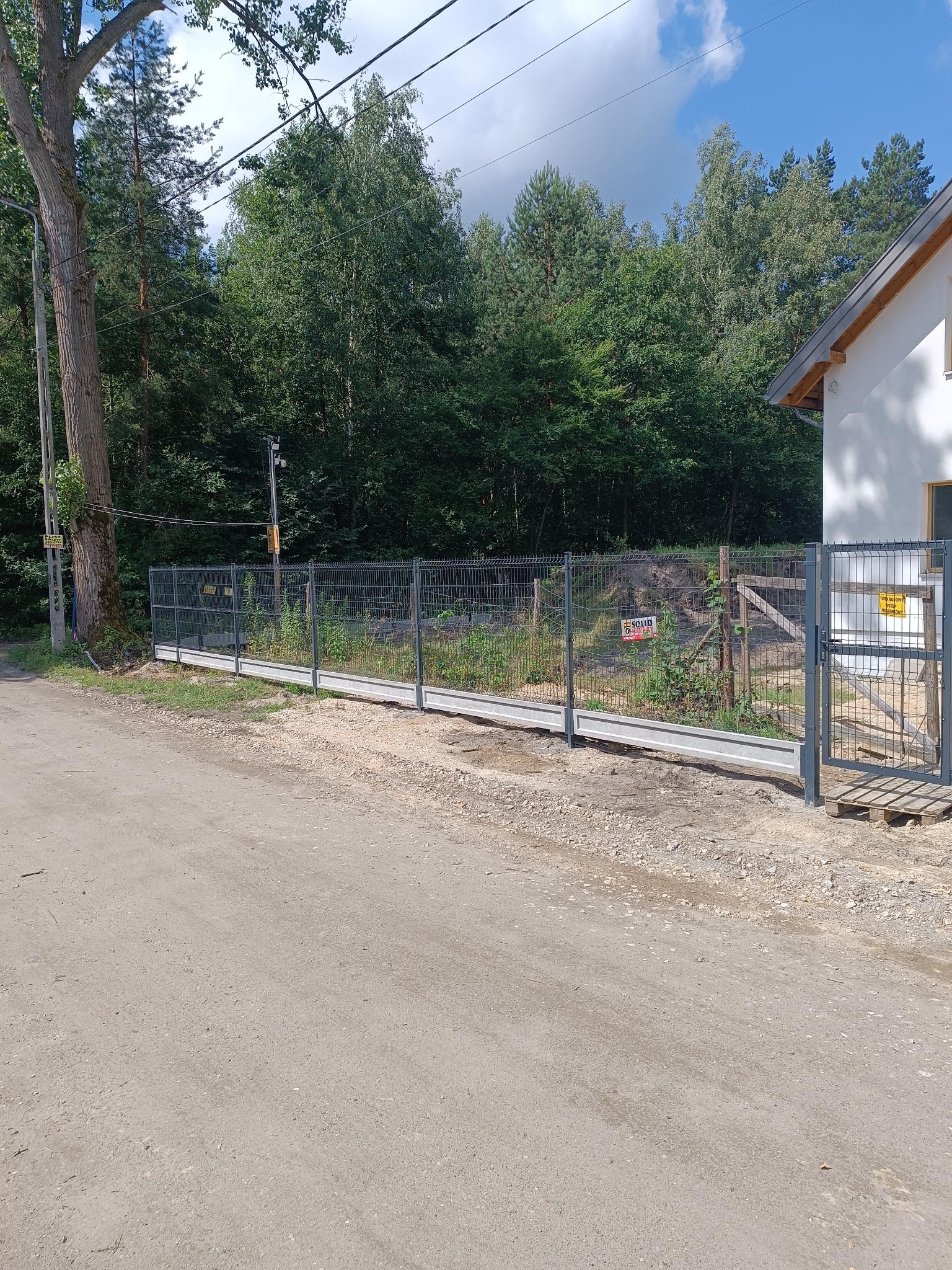 Szary panelowy płot z bramką na betonowej podmurówce, widok z zewnątrz posesji, w tle zieleń drzew i fragment białego budynku z szarym dachem.