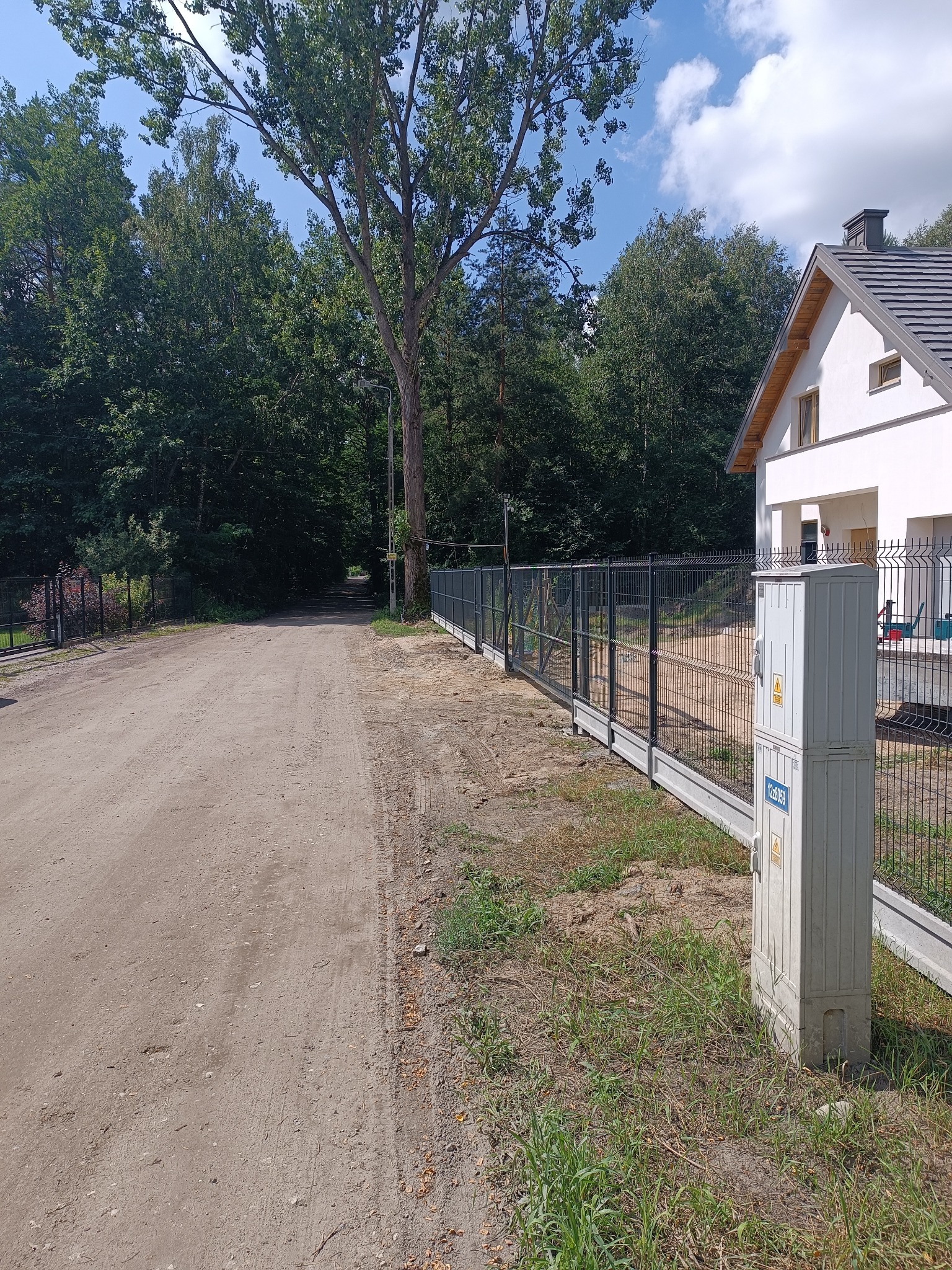 Nowoczesne, grafitowe ogrodzenie panelowe na betonowej podmurówce, z widoczną skrzynką elektryczną. Tło stanowi dom z białymi ścianami i zielone drzewa.