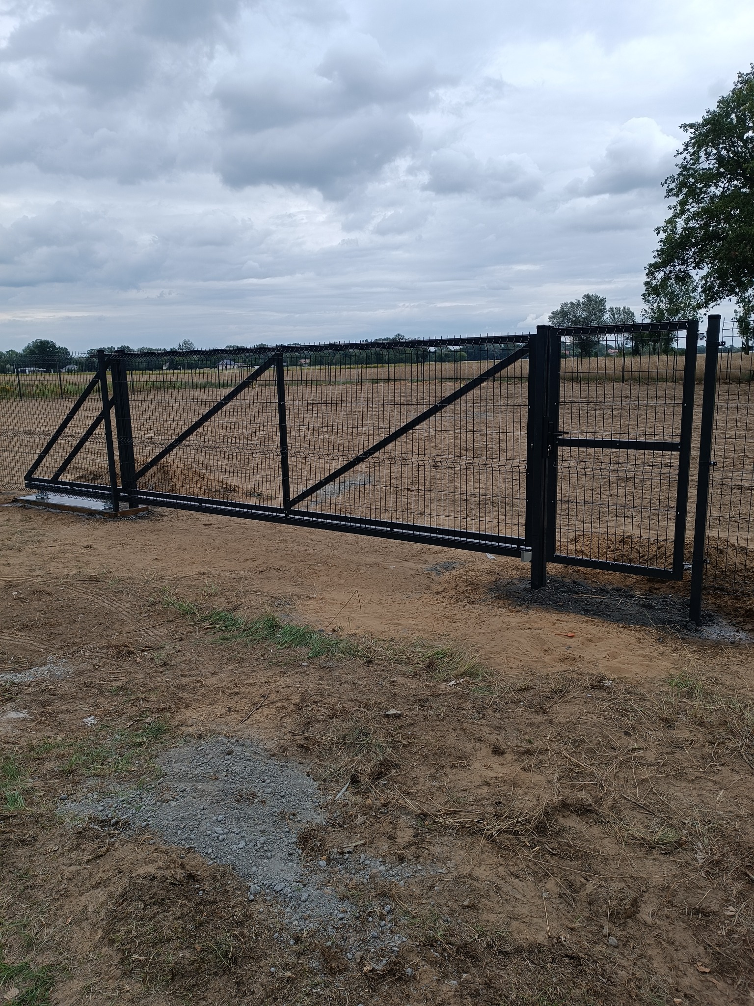 Czarna, przesuwna brama z siatki panelowej, zamontowana na posesji z widocznym gruntem i fragmentem drzewa w tle, pod pochmurnym niebem.