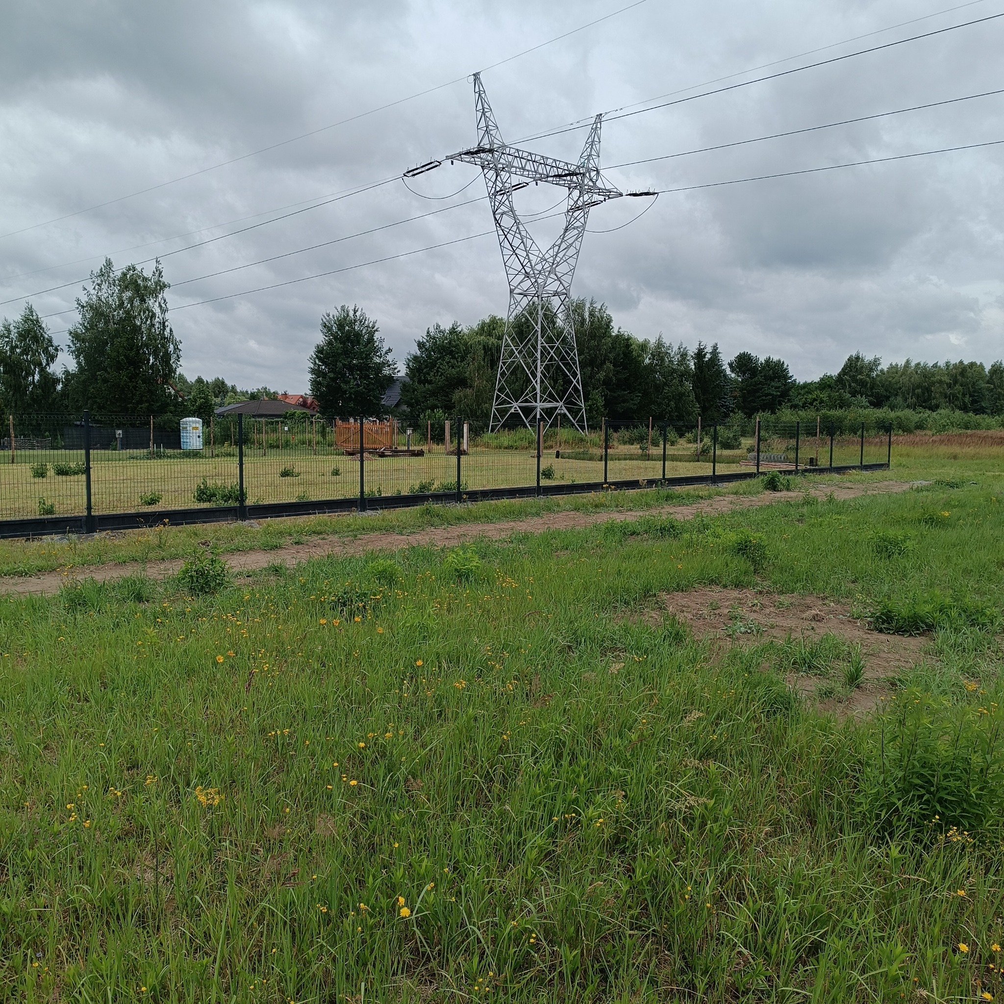 Czarny panelowy płot na tle łąki i słupa wysokiego napięcia. Widoczne drzewa i zabudowania w oddali. Pochmurne niebo.