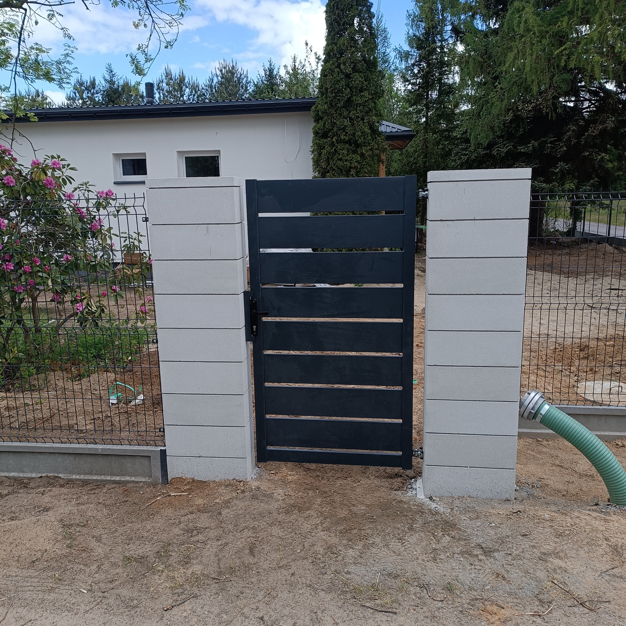 Nowoczesna furtka z poziomych profili, osadzona między słupkami z szarej kostki. W tle dom i ogrodzenie panelowe z siatki, obok furtki wąż ogrodowy.