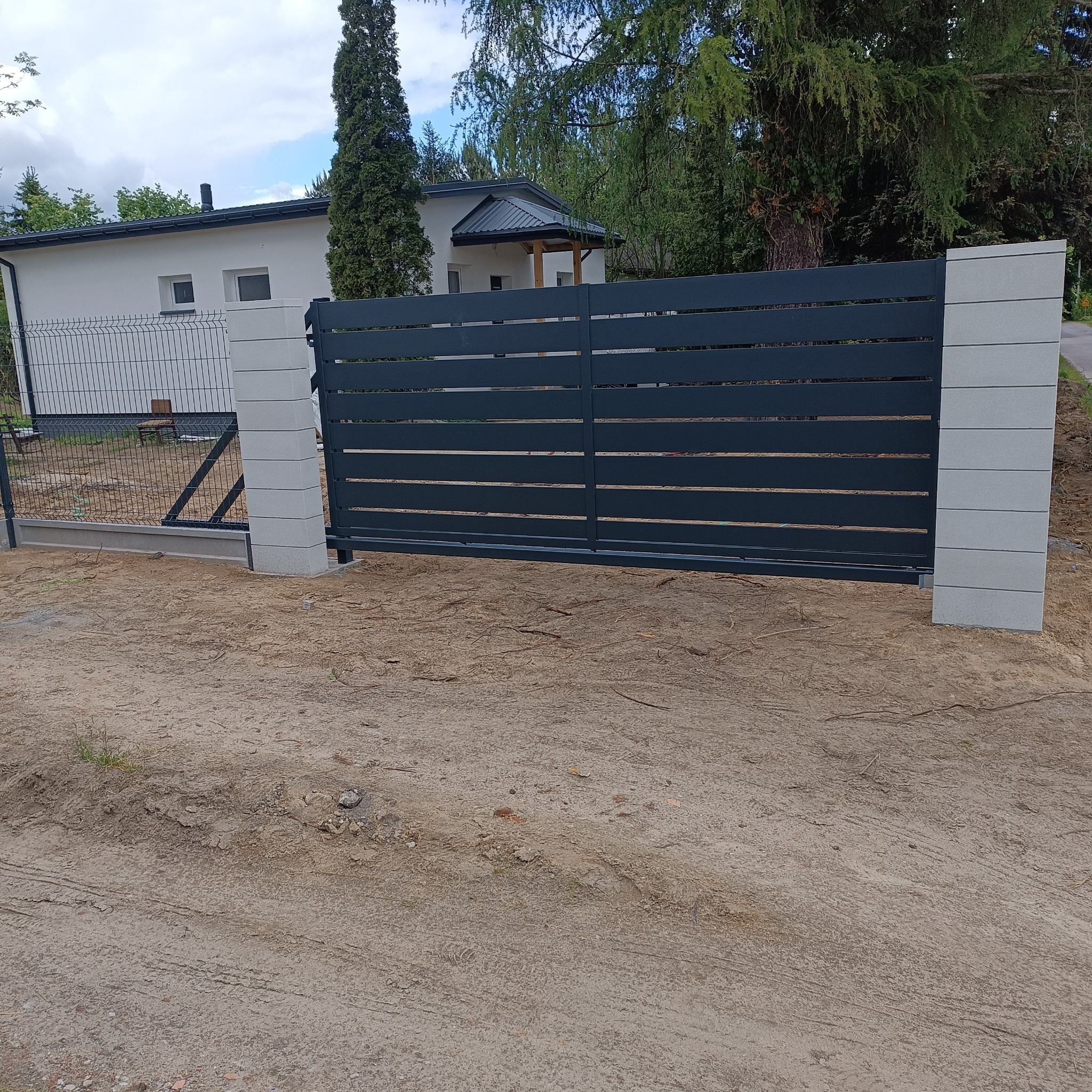 Nowoczesna, przesuwna brama w kolorze antracytu z poziomymi panelami, osadzona na betonowych słupkach. W tle dom z białymi ścianami i zielone drzewa.