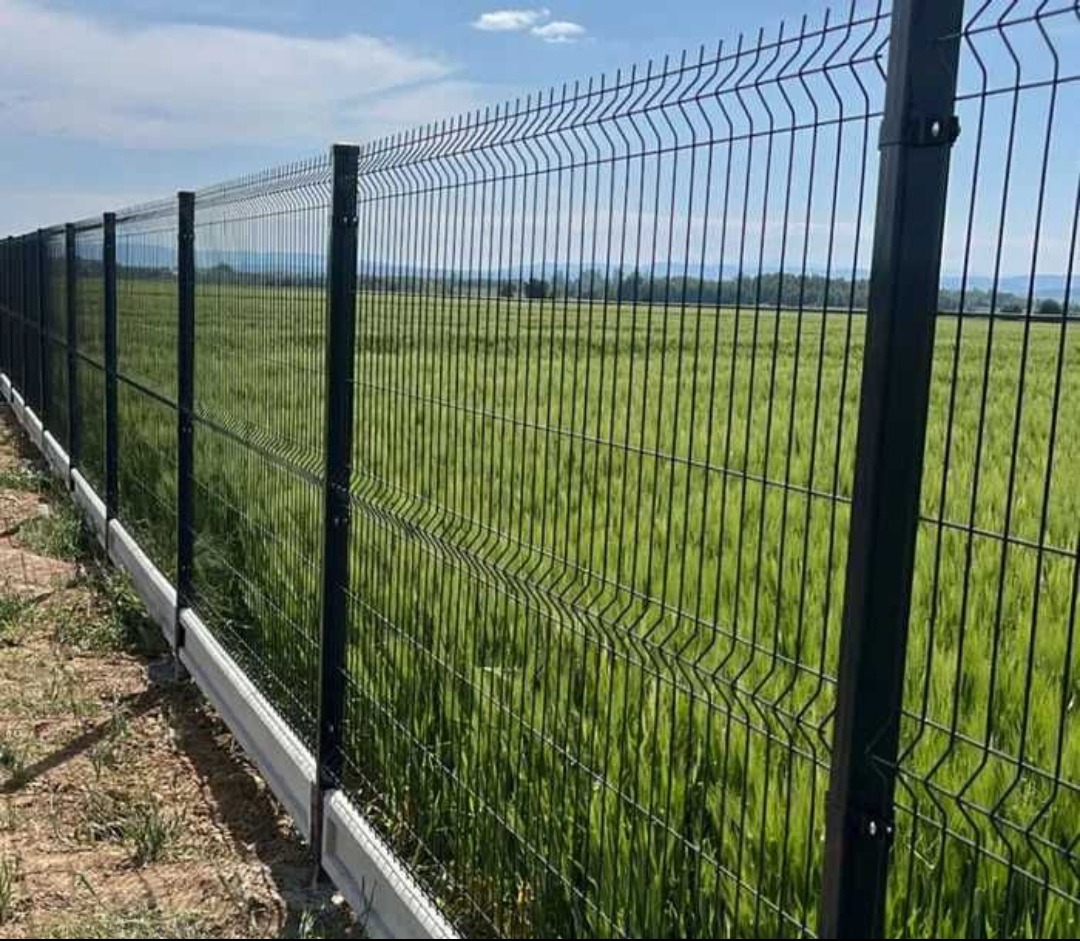Ciemne panele ogrodzeniowe z podmurówką na tle zielonego pola i błękitnego nieba. Widok z perspektywy, ukazujący długość ogrodzenia.