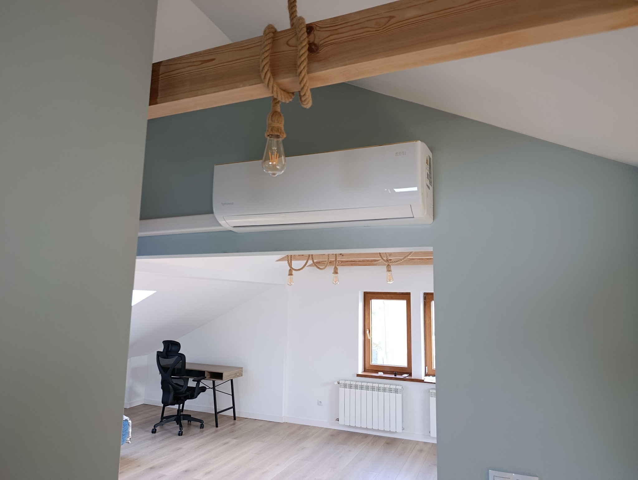 Biały klimatyzator Rotenso zamontowany na szarej ścianie pod drewnianą belką z dekoracyjną żarówką. Widok na jasne biuro z oknem i biurkiem.