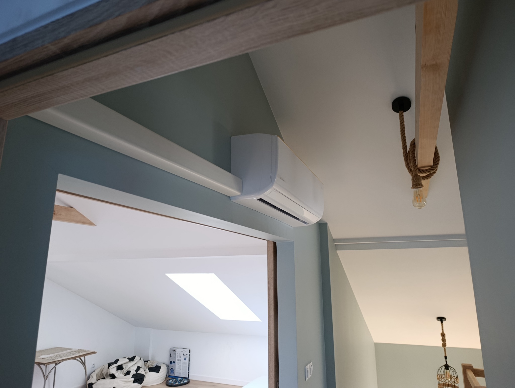 Biały klimatyzator zamontowany na ścianie w pokoju na poddaszu, widok z dołu. W tle widoczny fragment pokoju z oknem dachowym i dekoracyjną lampą.