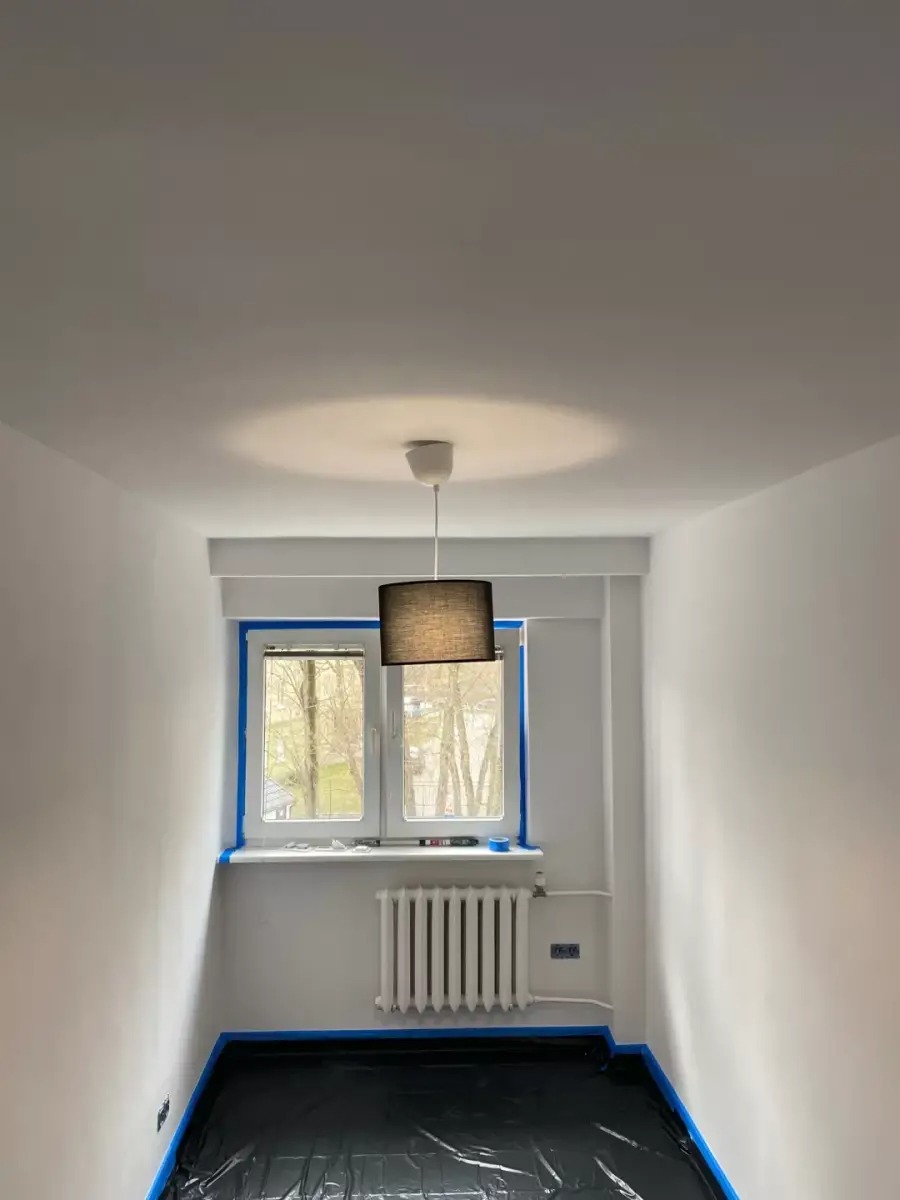 Pokój przygotowany do malowania: okno i podłoga zabezpieczone taśmą i folią, ściany świeżo pomalowane na biało, widoczny grzejnik i lampa.