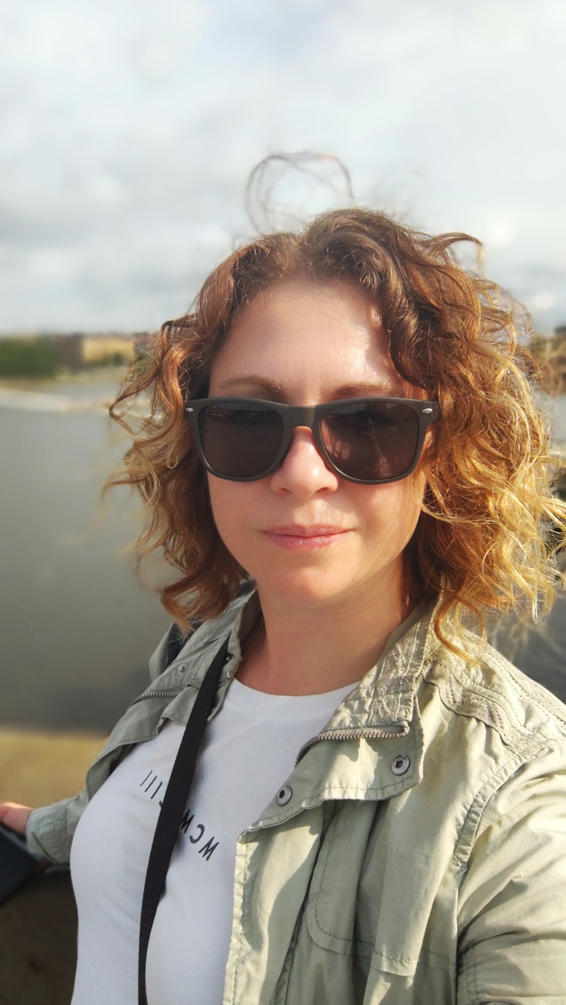 Portret kobiety w okularach przeciwsłonecznych na tle rzeki i krajobrazu miejskiego. Ubrana w kurtkę i białą koszulkę. Włosy kręcone, rude. Słoneczny dzień.