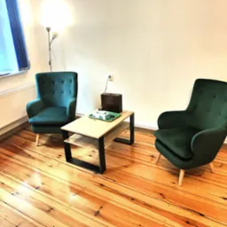 Jasne wnętrze gabinetu z dwoma zielonymi fotelami i stolikiem kawowym. Na stoliku pudełko herbaty Vertea. Drewniana podłoga i białe ściany. Przytulna atmosfera.