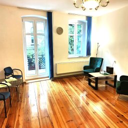 Wielkopolski Instytut Neurolingwistyki Centrum Psychoterapii - Jasne wnętrze gabinetu z drewnianą podłogą, zielonymi fotelami, stolikiem i oknami z widokiem na zieleń. Na ścianie zegar.
