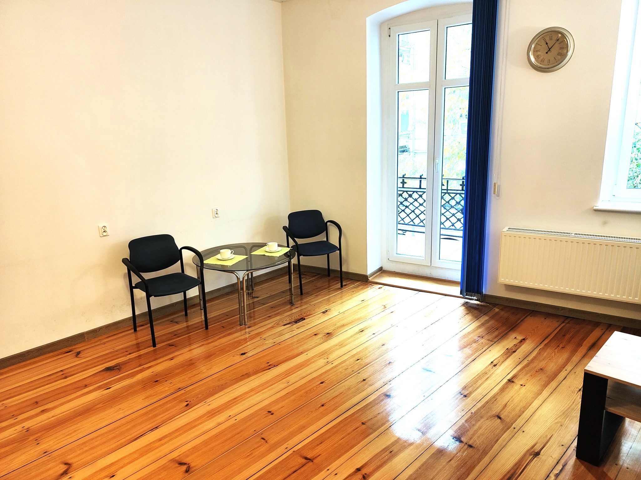 Jasne wnętrze gabinetu z drewnianą podłogą, dwoma krzesłami i stolikiem. Na ścianie wisi zegar, a przez okno widać zieleń. Pokój przygotowany do spotkania.