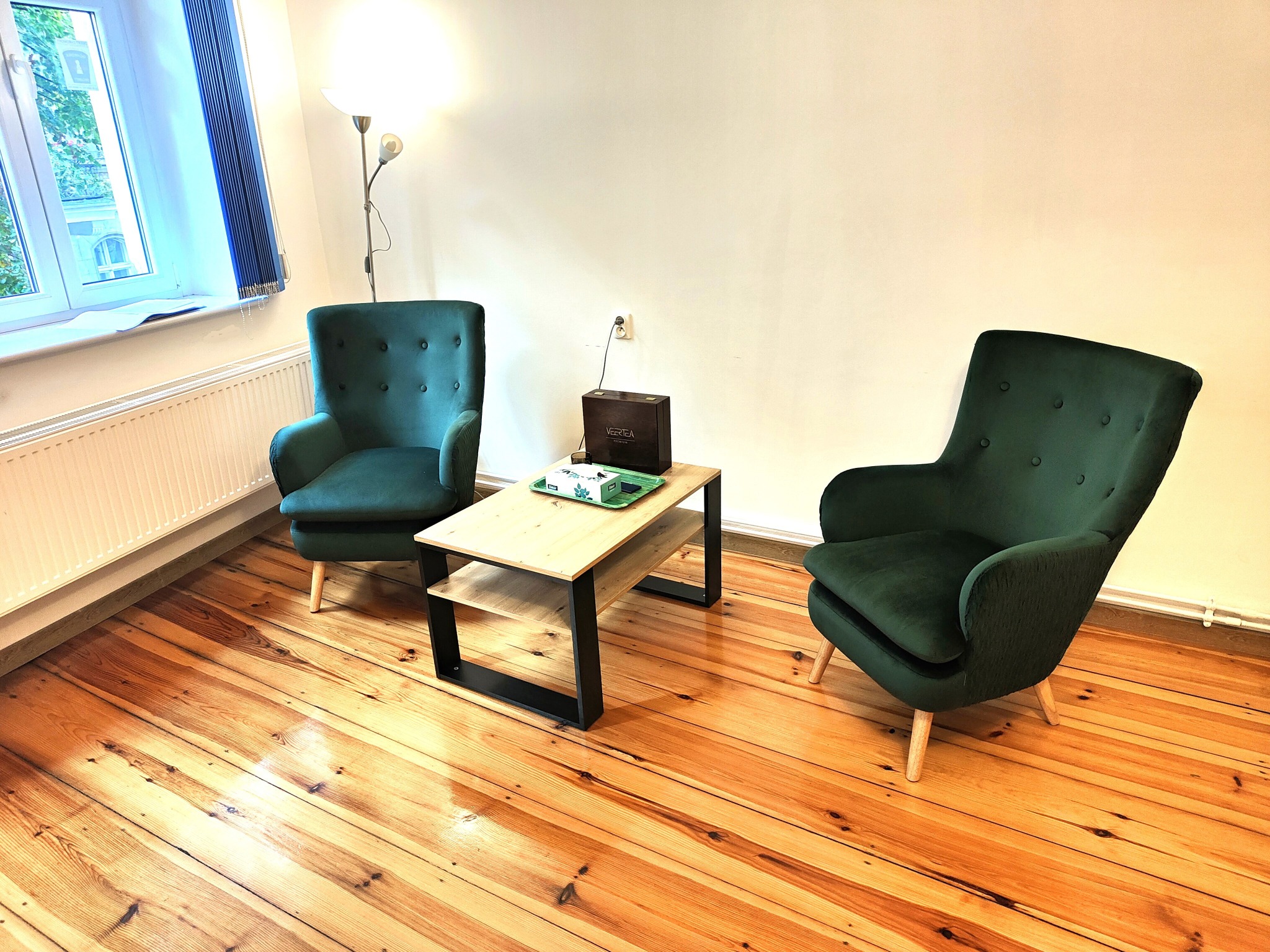 Jasne wnętrze gabinetu z dwoma zielonymi fotelami i stolikiem kawowym. Na stoliku pudełko herbaty Vertea. Drewniana podłoga i białe ściany. Przytulna atmosfera.