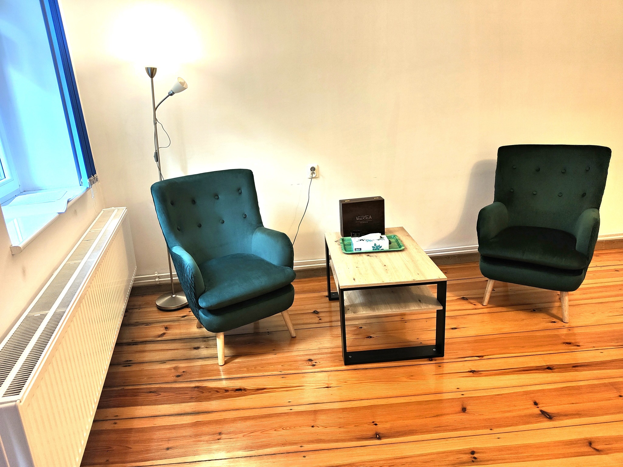 Dwa zielone fotele klubowe ustawione w jasnym pokoju na drewnianej podłodze, stolik kawowy z pudełkiem herbaty i chusteczkami, lampa podłogowa i okno z zasłoną.