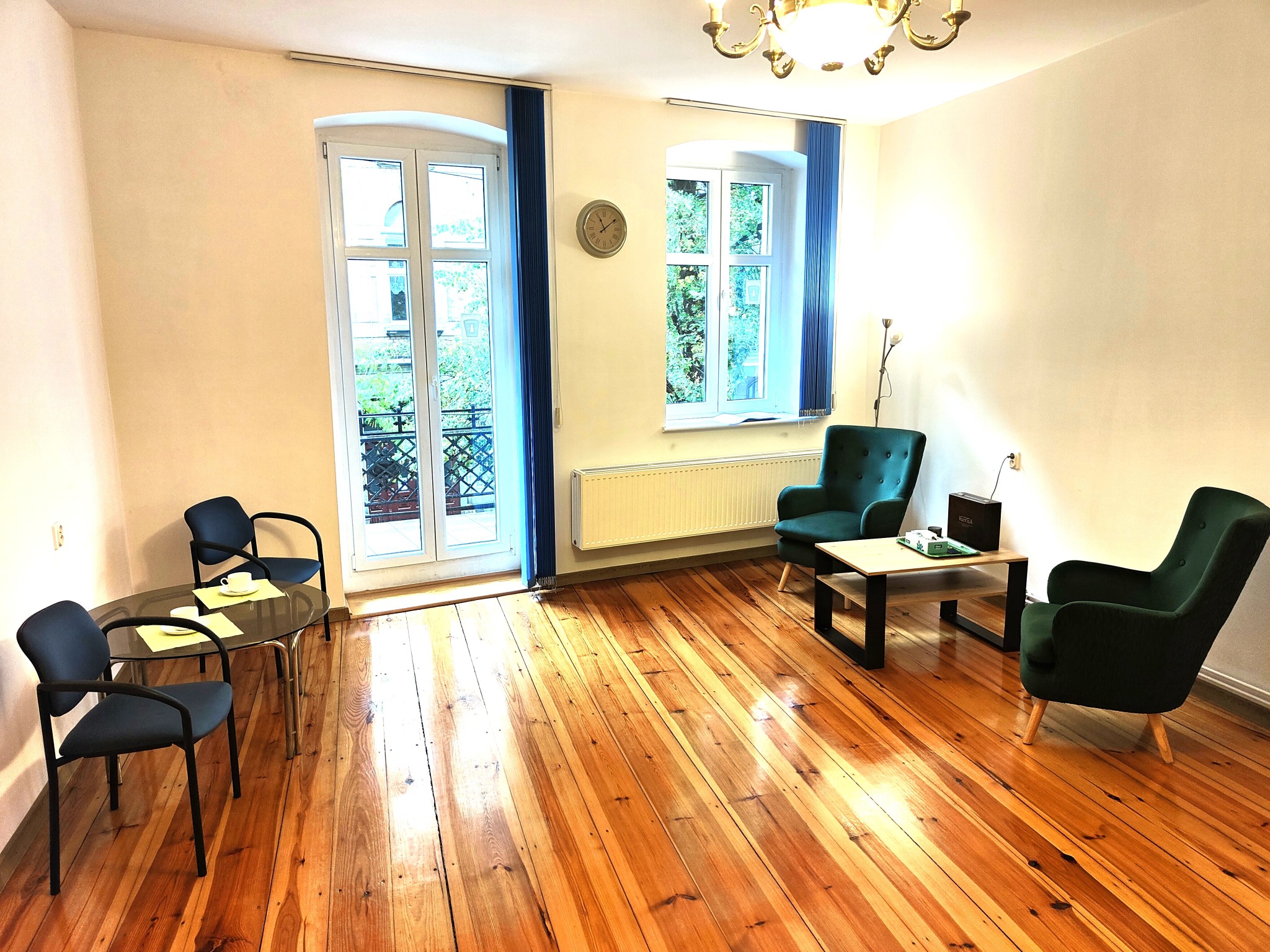 Jasne wnętrze gabinetu z drewnianą podłogą, zielonymi fotelami, stolikiem i oknami z widokiem na zieleń. Na ścianie zegar.