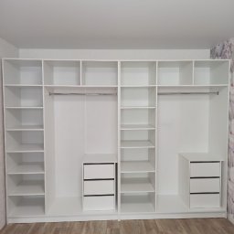 Vladyslav Lebid - Biała, zabudowana szafa z szufladami i półkami, widok z przodu. Minimalistyczny design, jasne wnętrze, drewnopodobna podłoga i tapeta w kwiaty w tle.