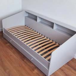 Vladyslav Lebid - Szare łóżko z szufladami i półkami wbudowanymi w wezgłowie, widoczne drewniane listwy stelaża, na podłodze panele imitujące drewno, minimalistyczny design.