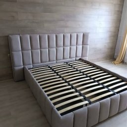 Vladyslav Lebid - Tapicerowane łóżko z wysokim wezgłowiem w odcieniach szarości, z drewnianym stelażem, na tle ściany z paneli i delikatnej firanki.