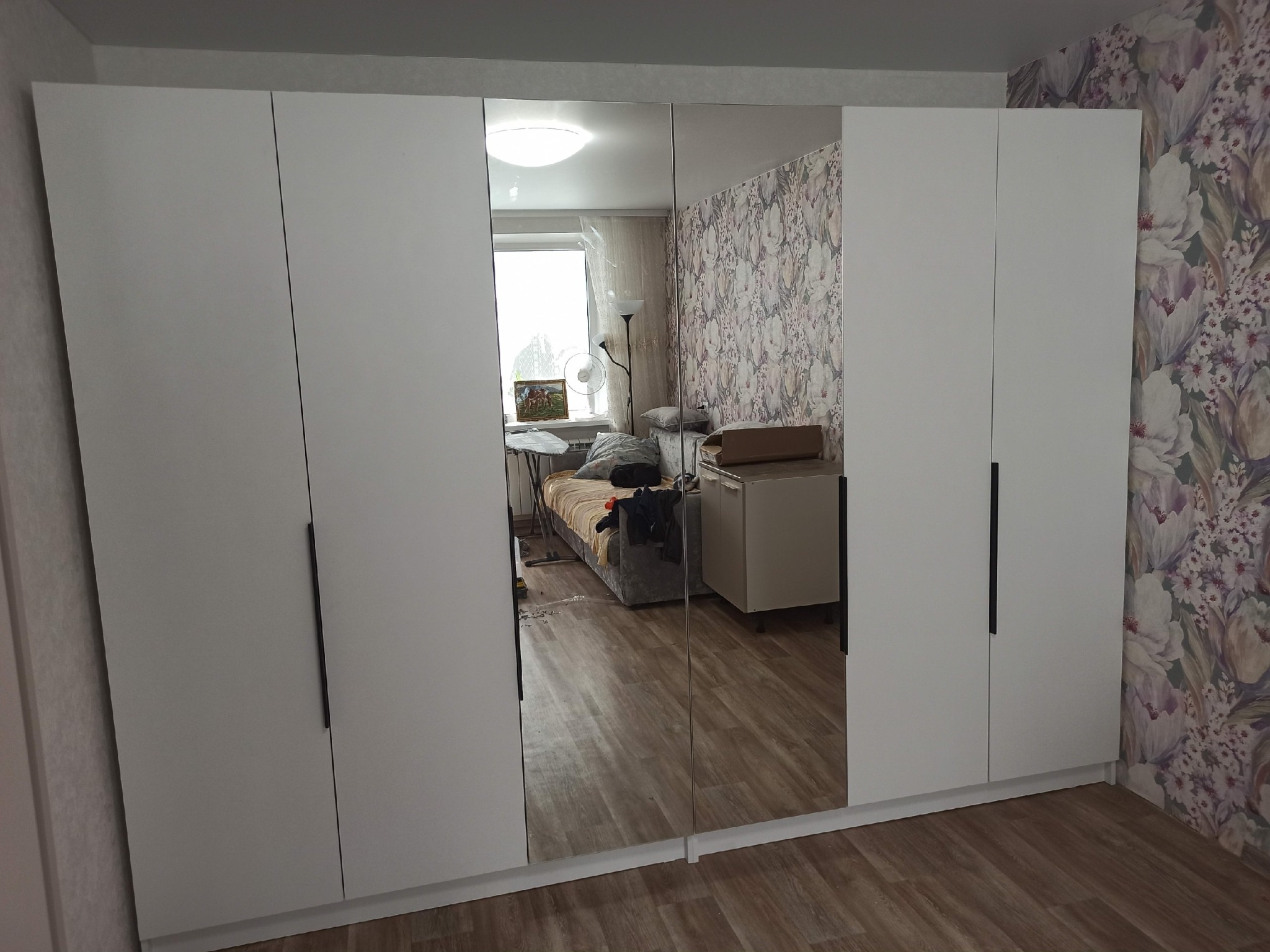 Duża, biała szafa z lustrzanymi drzwiami, odbijająca wnętrze pokoju z wzorzystą tapetą i elementami wyposażenia. Minimalistyczny design, proste uchwyty.