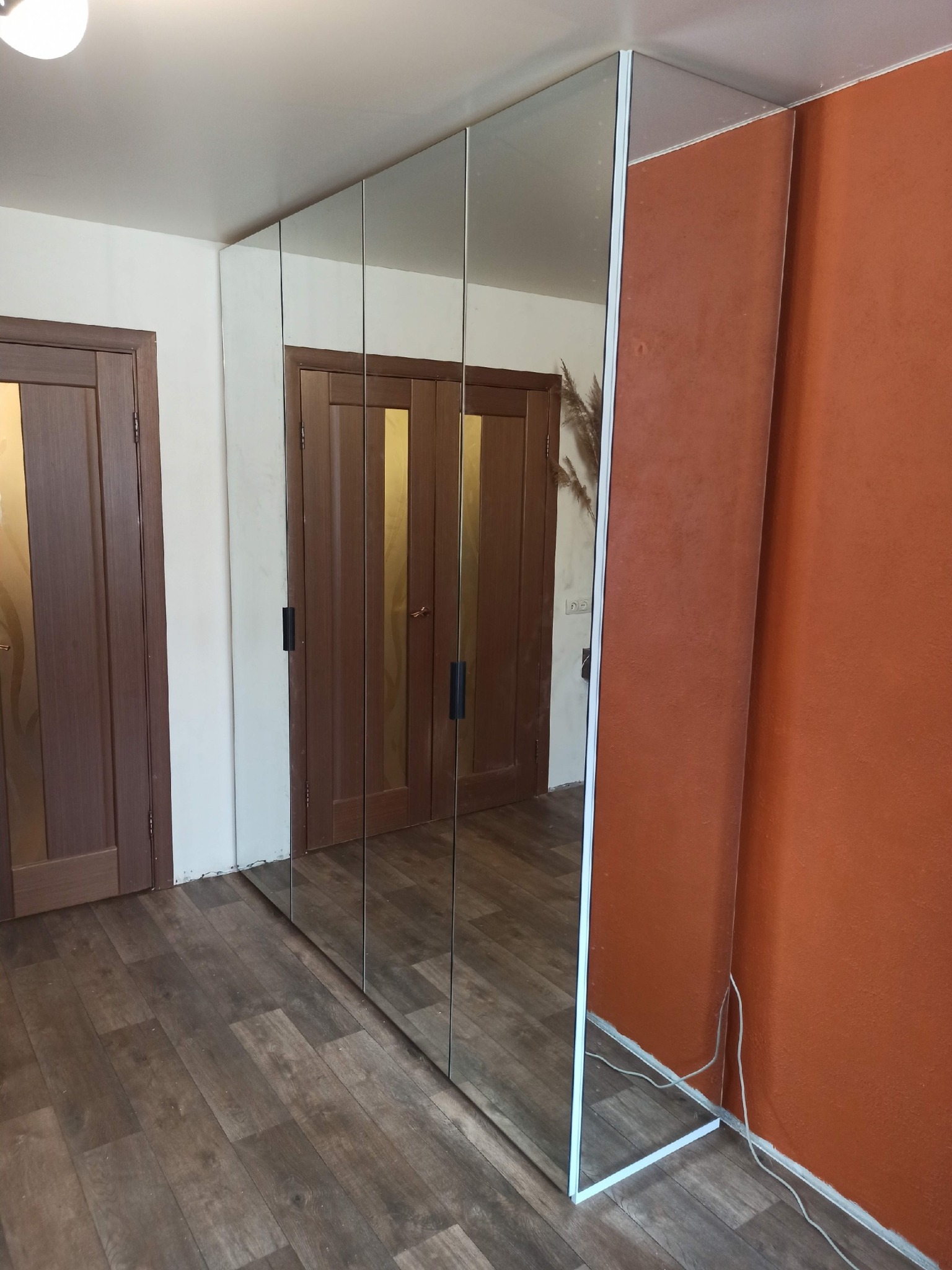 Nowoczesna szafa wnękowa z lustrzanymi drzwiami, wkomponowana w przestrzeń między drzwiami i ścianą w odcieniach brązu. Widoczne panele podłogowe.