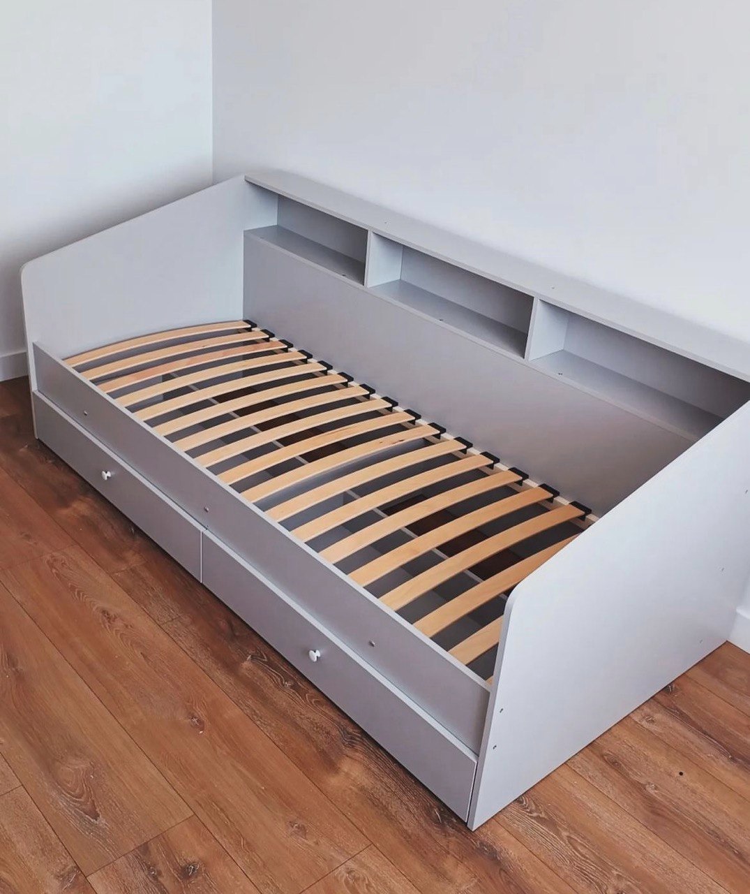 Szare łóżko z szufladami i półkami wbudowanymi w wezgłowie, widoczne drewniane listwy stelaża, na podłodze panele imitujące drewno, minimalistyczny design.