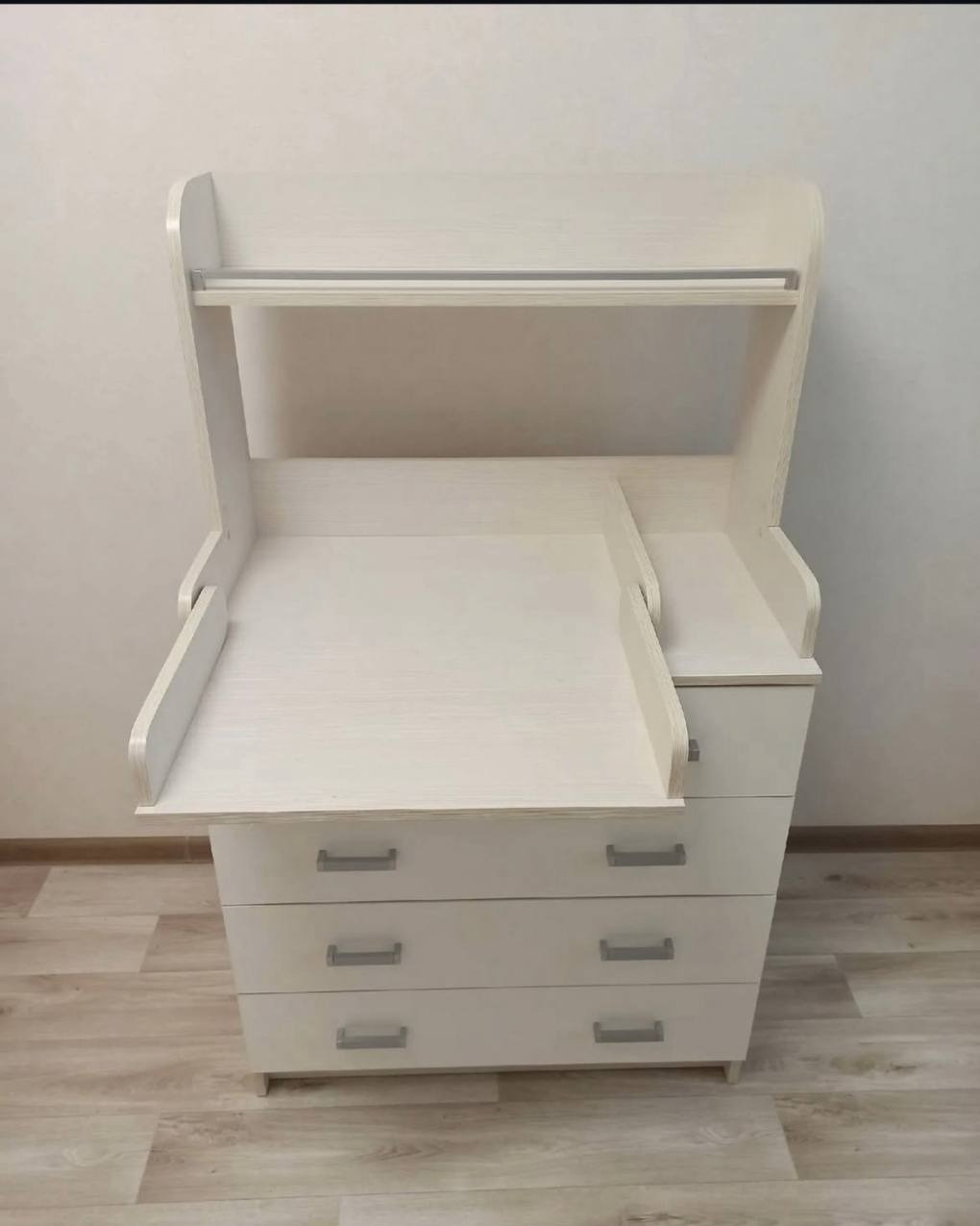 Biała komoda z przewijakiem i trzema szufladami, widok z przodu na tle jasnej ściany i podłogi z paneli. Przewijak rozłożony po lewej stronie.