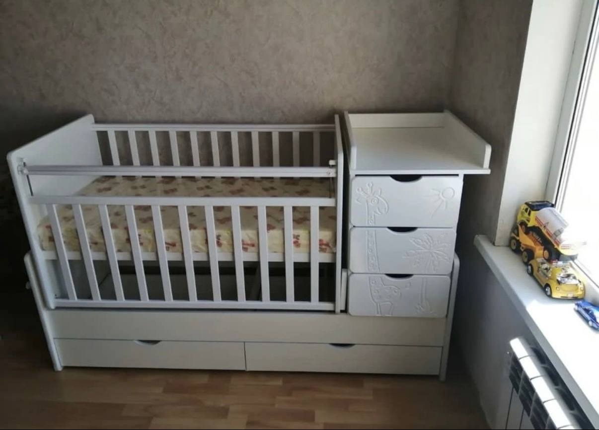 Białe łóżeczko dziecięce z przewijakiem i szufladami, zdobione motywami zwierząt i roślin. Obok okno z zabawkami. Całość w jasnym, neutralnym wnętrzu.