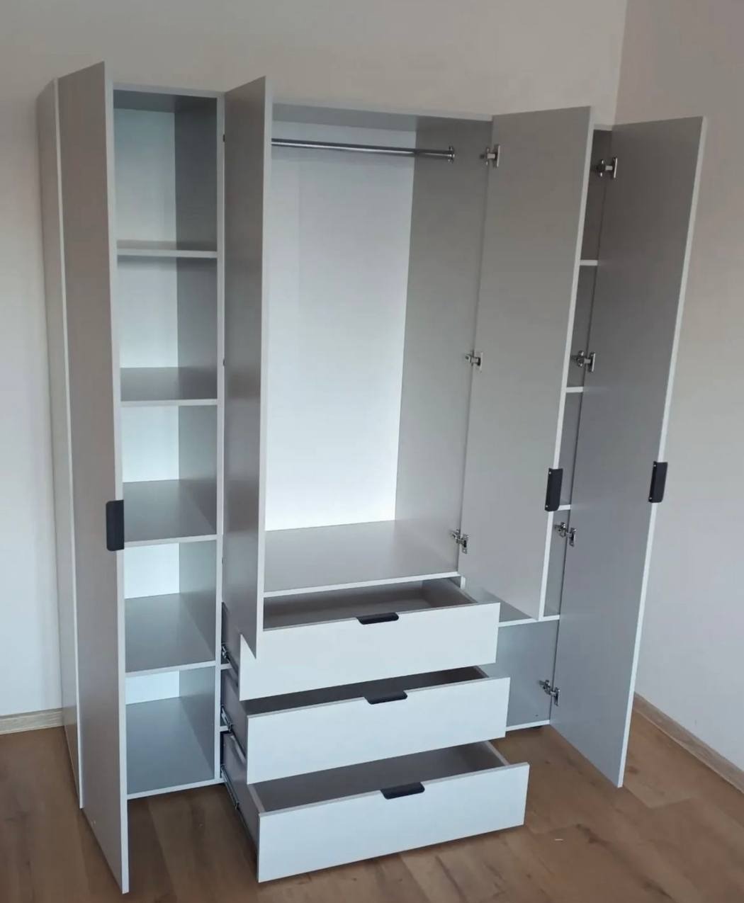 Nowoczesna, biała szafa z otwartymi drzwiami i wysuwanymi szufladami, widok z przodu. Montaż mebli w Warszawie, widoczne półki i drążek na ubrania.