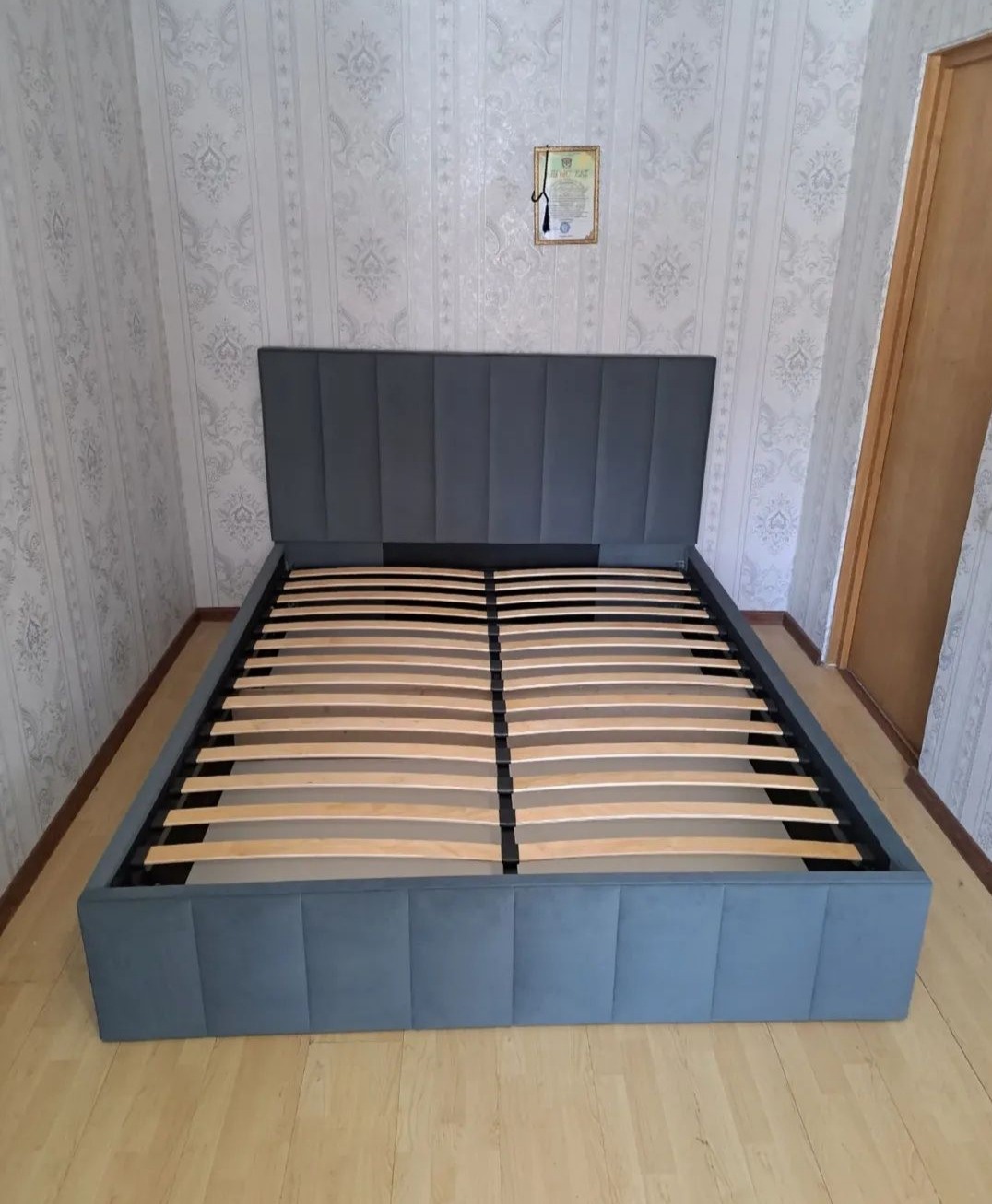 Tapicerowane łóżko z drewnianym stelażem w pokoju w Warszawie. Szary zagłówek z pionowymi przeszyciami. Widok z przodu, neutralne oświetlenie.