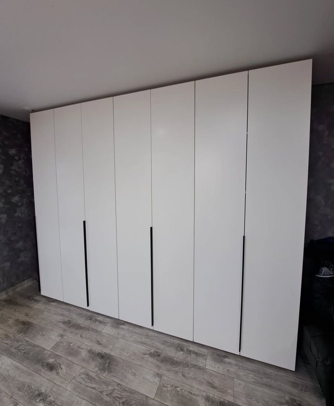 Biała szafa wnękowa z czarnymi, minimalistycznymi uchwytami, dopasowana do szarej podłogi i ściany z efektem dekoracyjnym. Nowoczesny design.