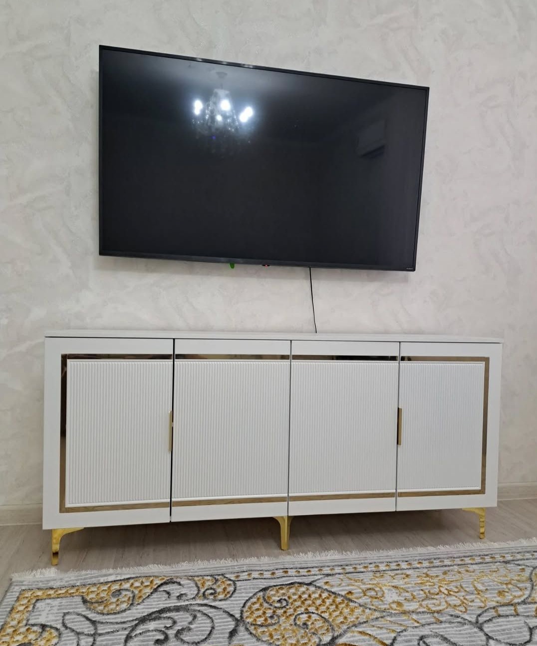 Biała szafka RTV z ryflowanymi drzwiczkami i złotymi akcentami, ustawiona na tle wzorzystej ściany z zawieszonym telewizorem. Widoczny fragment dywanu.
