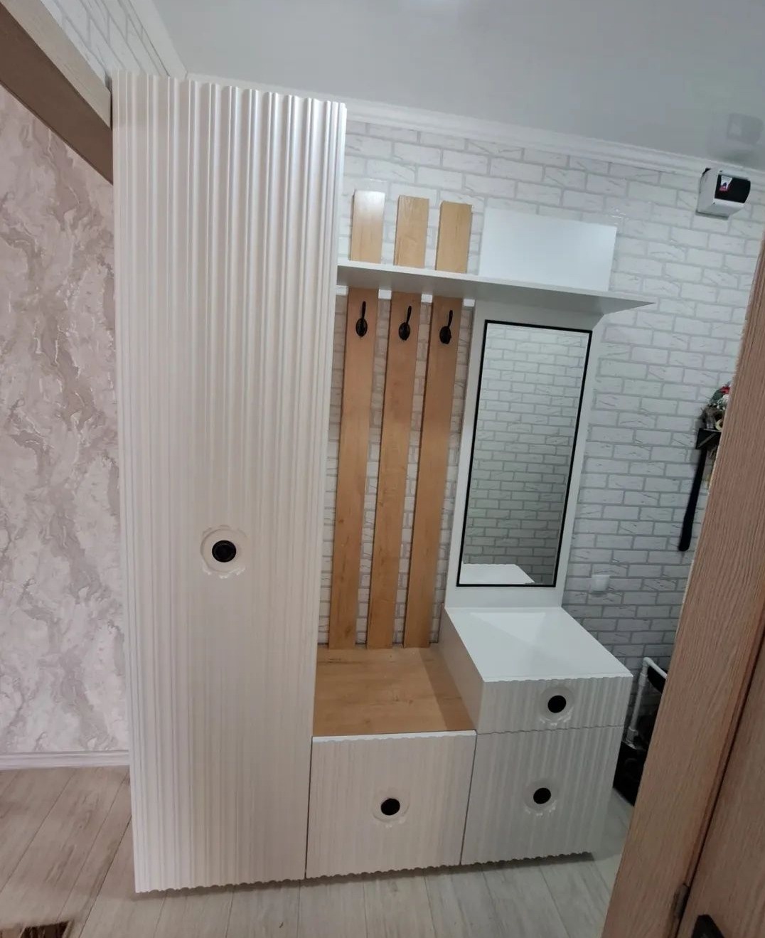 Biały, nowoczesny przedpokój z szafą, lustrem i wieszakami na tle ceglanej ściany. Charakterystyczne okrągłe uchwyty.