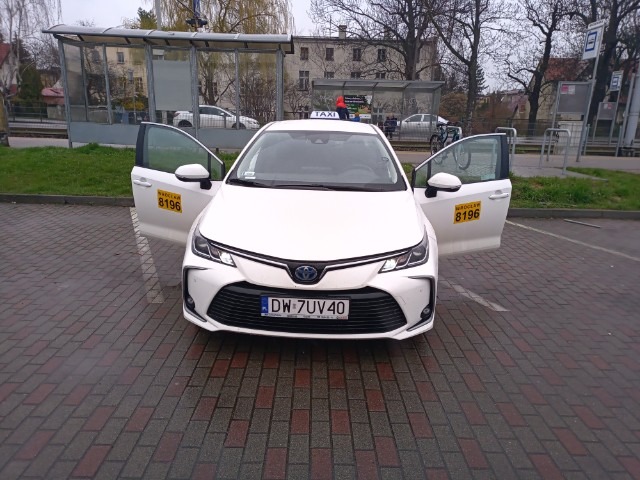 Biała taksówka Toyota Corolla z otwartymi drzwiami na parkingu, widok z przodu. Numer licencji i oznaczenie taxi na dachu. Przystanek autobusowy w tle.