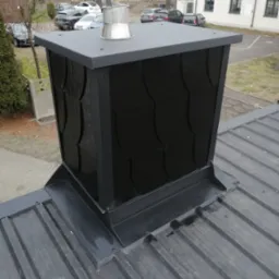 Szary, metalowy komin z dekoracyjnym wykończeniem na dachu z blachy trapezowej. Widoczne obróbki blacharskie i nasada kominowa. Tło stanowią budynki i drzewa.