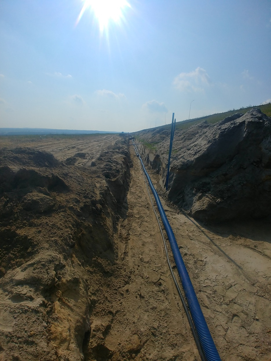 Wykop z niebieską rurą karbowaną i kablem na terenie budowy KGHM Rudna. Instalacja podziemna w piaszczystym gruncie, widok z góry na wykop.