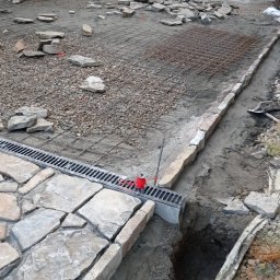 PAULAMIR PAULINA LISZKA - Układanie kamiennej nawierzchni z odpływem liniowym. Widoczna siatka zbrojeniowa, kamienie do ułożenia i wykop. Prace brukarskie w toku.