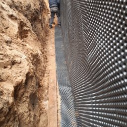 PAULAMIR PAULINA LISZKA - Prace ziemne przy fundamencie z widoczną folią kubełkową. Pracownik w trakcie montażu. Wykop zabezpieczony deską. Grunt piaszczysty.