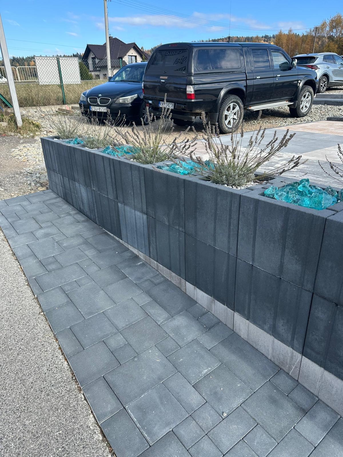 Mur z betonowych bloczków z roślinami i szkłem dekoracyjnym, obok ułożona kostka brukowa. W tle zaparkowane samochody i dom.