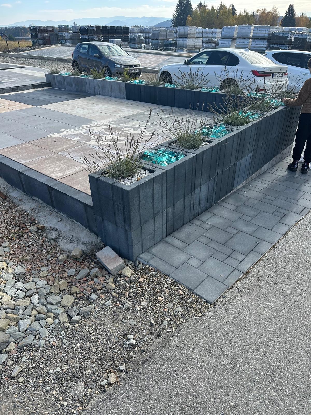 Aranżacja przestrzeni z kostki brukowej i betonowych donic z roślinami oraz dekoracyjnym szkłem. Widoczne samochody i palety z kostką w tle.