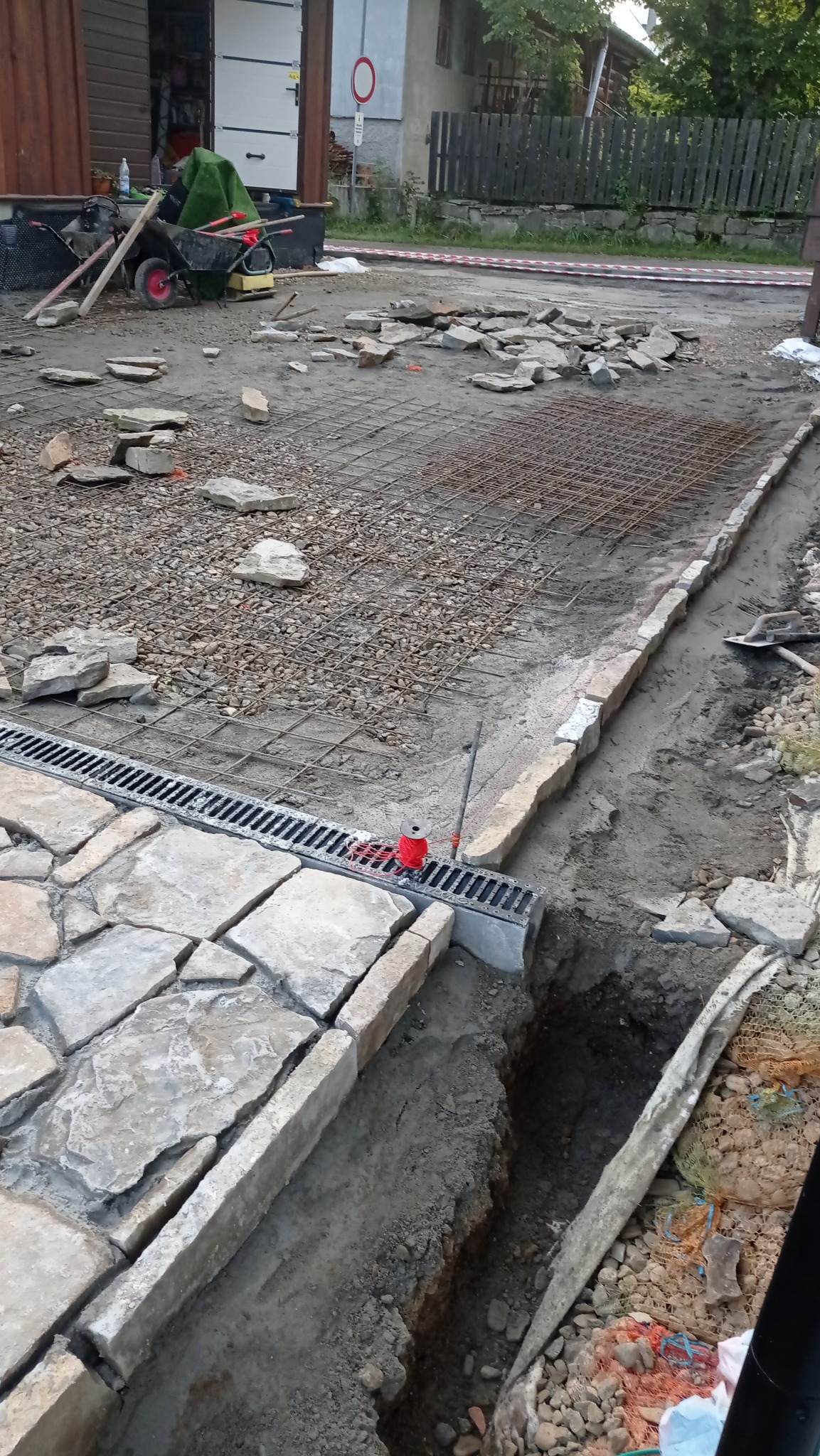 Układanie kamiennej nawierzchni z odpływem liniowym. Widoczna siatka zbrojeniowa, kamienie do ułożenia i wykop. Prace brukarskie w toku.