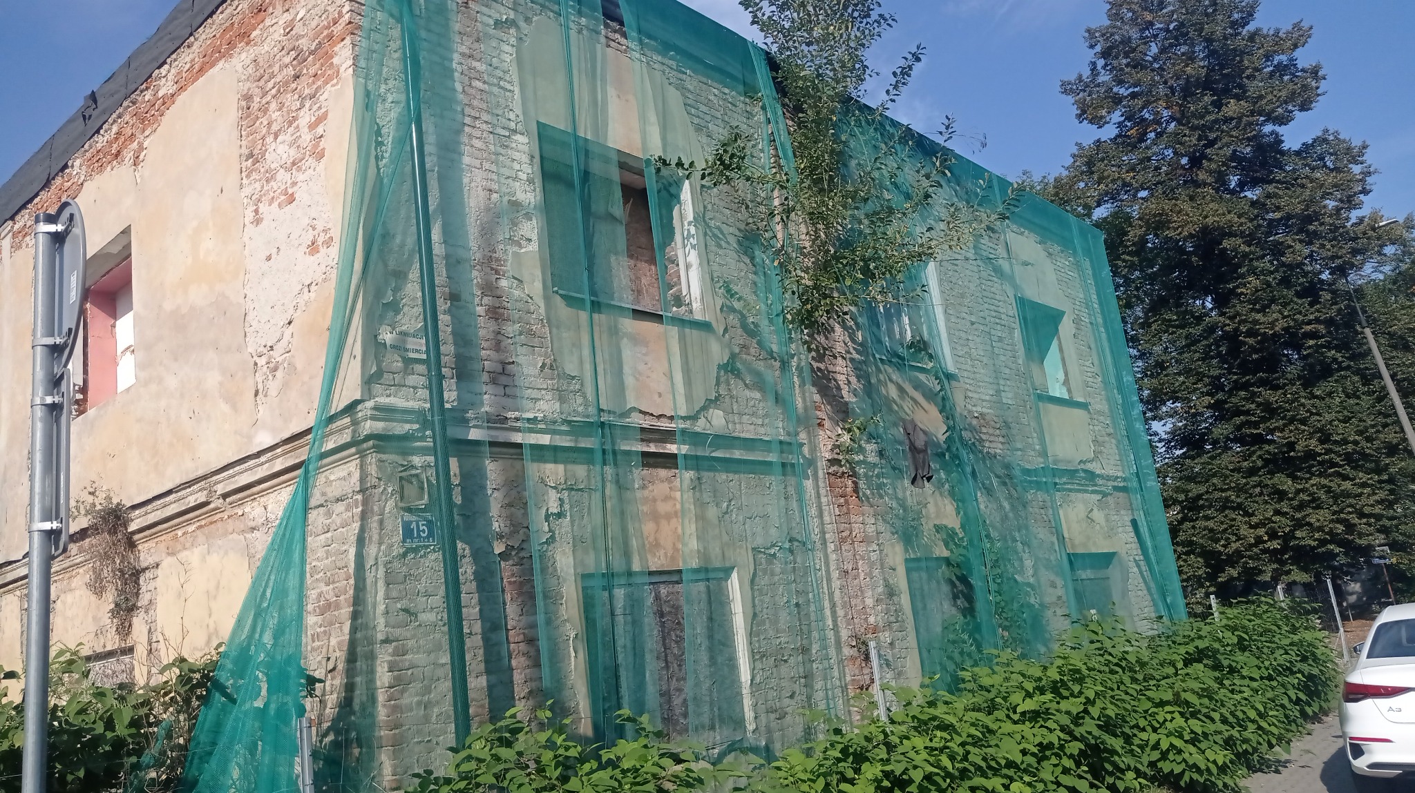 Stara, ceglana kamienica w trakcie rozbiórki, zabezpieczona zieloną siatką ochronną. Widoczne fragmenty elewacji, okna i zieleń. Ujęcie z dołu, w tle drzewa i fragment samochodu.