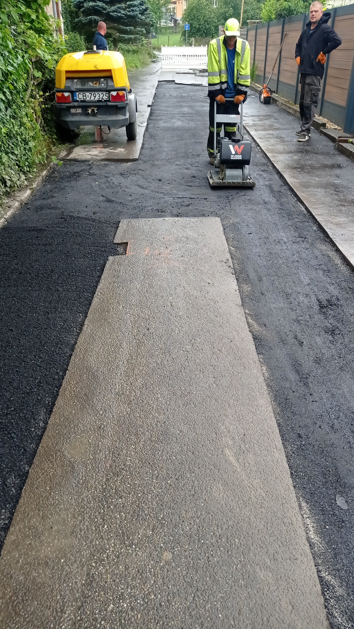 Pracownik w odblaskowej kamizelce zagęszcza asfaltową nawierzchnię ubijarką. Widoczny fragment nowej drogi, maszyna i ekipa remontowa. Praca w toku.