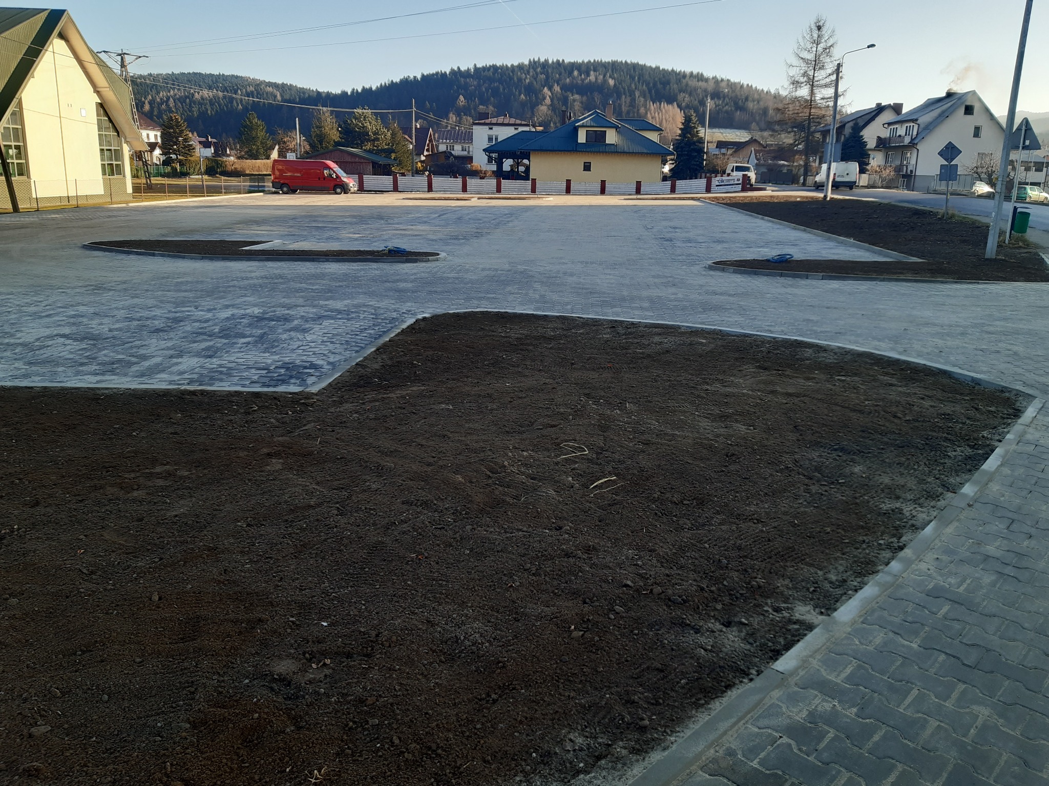 Nowa nawierzchnia z kostki brukowej na parkingu, z przygotowanymi rabatami ziemnymi. W tle budynki mieszkalne i zalesione wzgórze w słoneczny dzień.