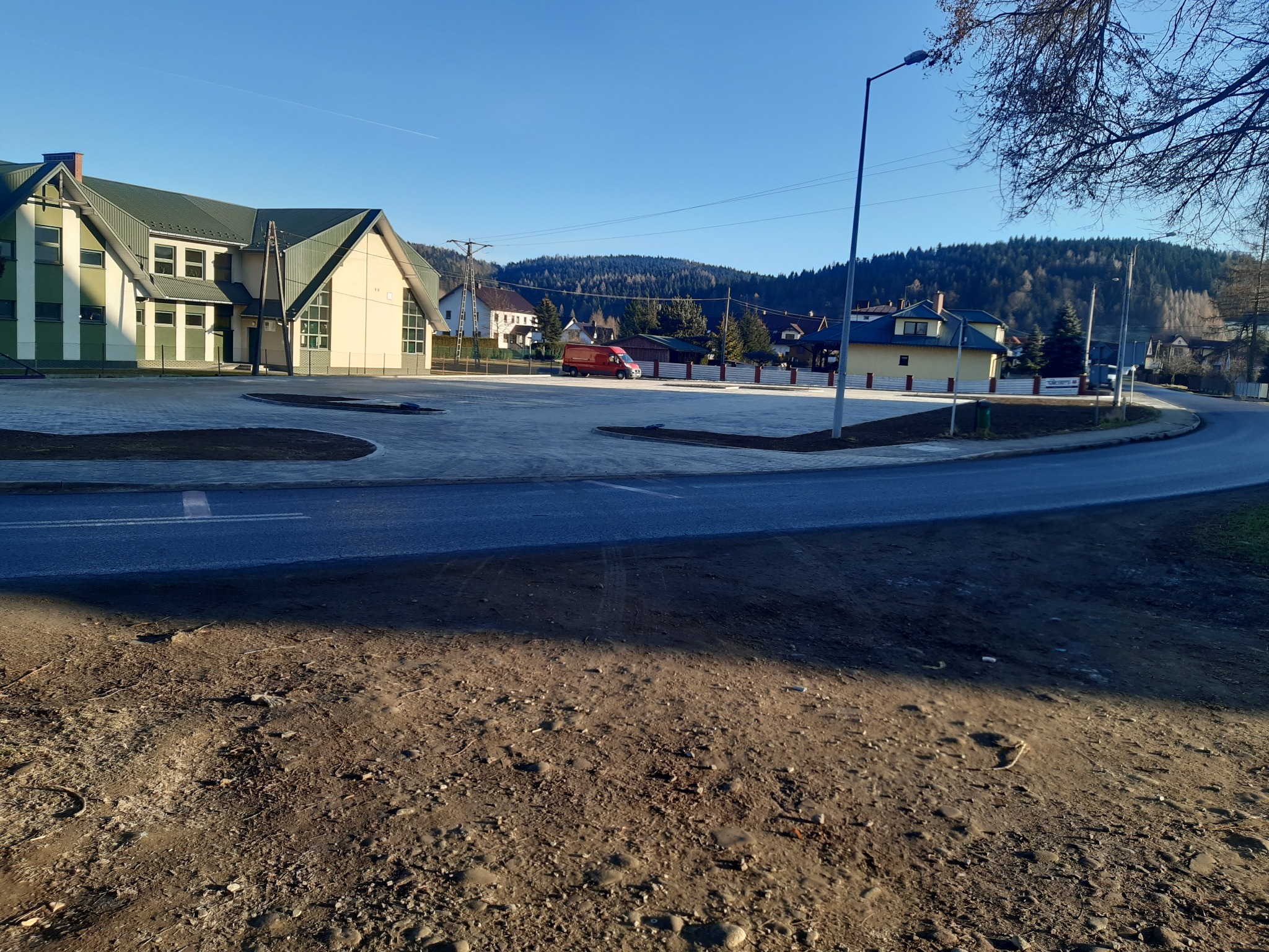 Rozległy plac z nową nawierzchnią z kostki brukowej przed budynkiem w jasnych barwach, w tle zalesione wzgórza i błękitne niebo. Widok z poziomu gruntu.