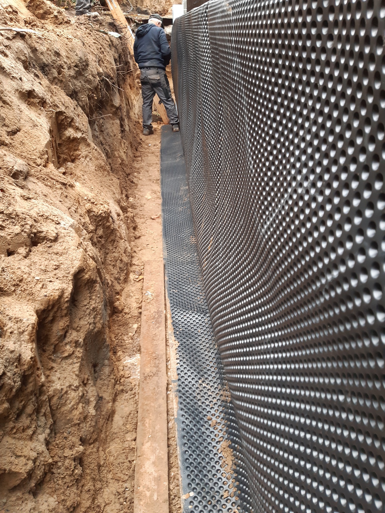Ochrona fundamentów: Czarna folia kubełkowa z otworami, zabezpieczająca wykopany fundament. Widoczny pracownik w wykopie. Grunt w kolorze beżowo-brązowym.