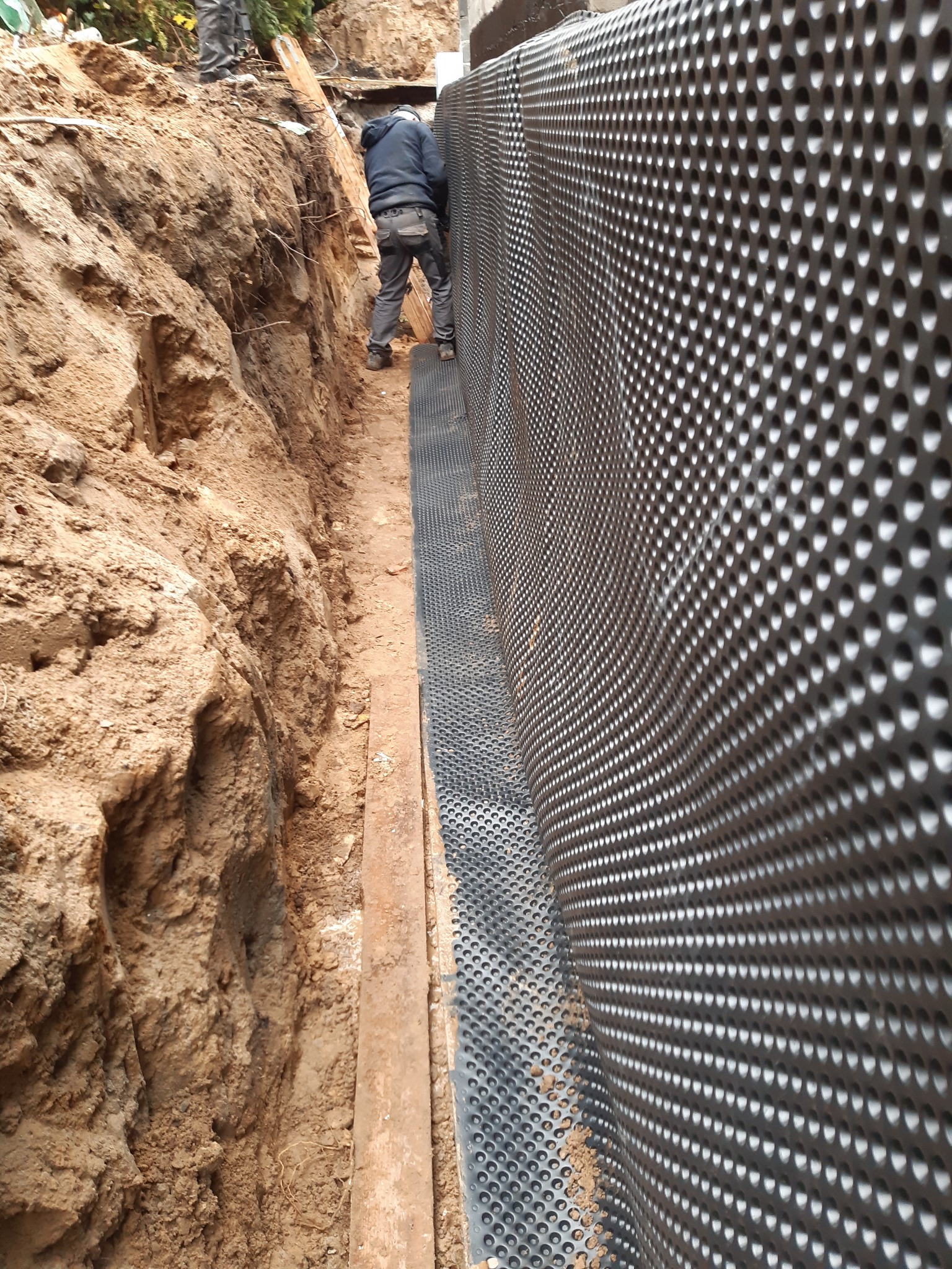 Prace ziemne przy fundamencie z widoczną folią kubełkową. Pracownik w trakcie montażu. Wykop zabezpieczony deską. Grunt piaszczysty.