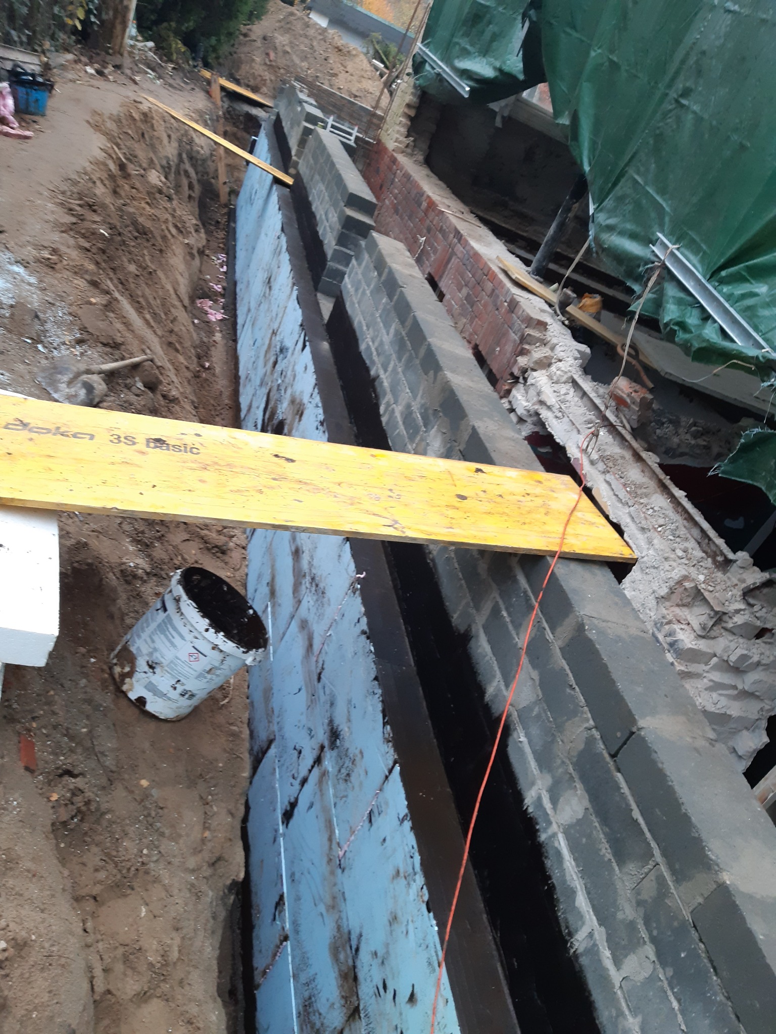 Fundamenty z izolacją termiczną w trakcie budowy, widoczne bloczki fundamentowe, styropian i żółta deska szalunkowa. Wykop ziemny i wiadro obok.