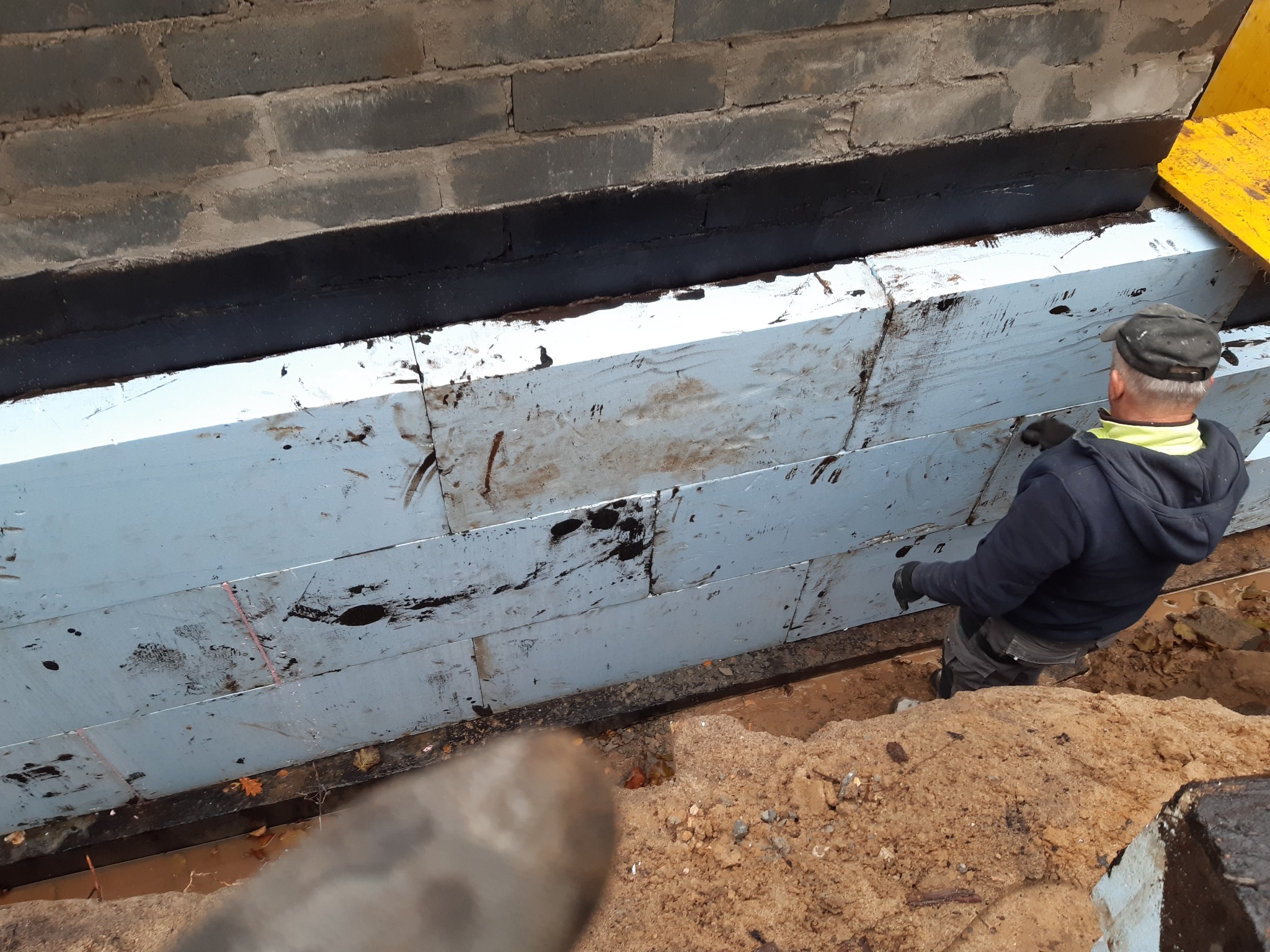 Ocieplenie fundamentów styropianem grafitowym, zabezpieczone czarną izolacją. Widoczny fragment ściany fundamentowej i pracownik w trakcie montażu.